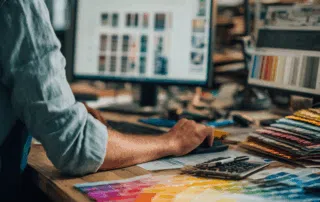 Graphiste travaillant sur un ordinateur avec des nuanciers de couleurs étalés sur un bureau en atelier